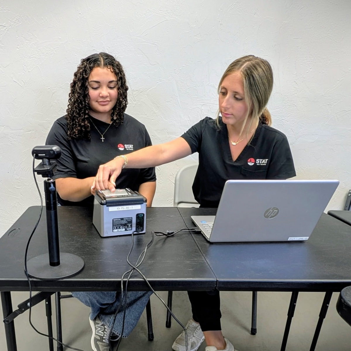 Two women working together at a table with a laptop and other equipment.